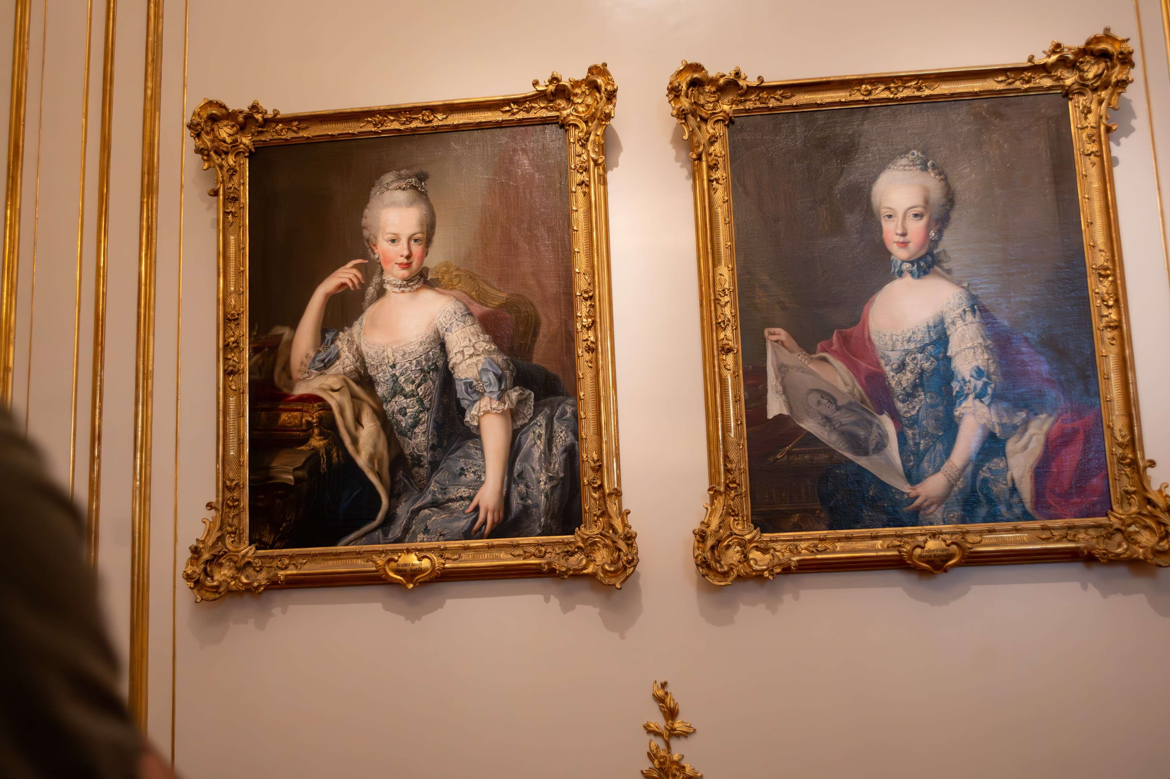 Two paintings of women in formal dresses are on a wall.