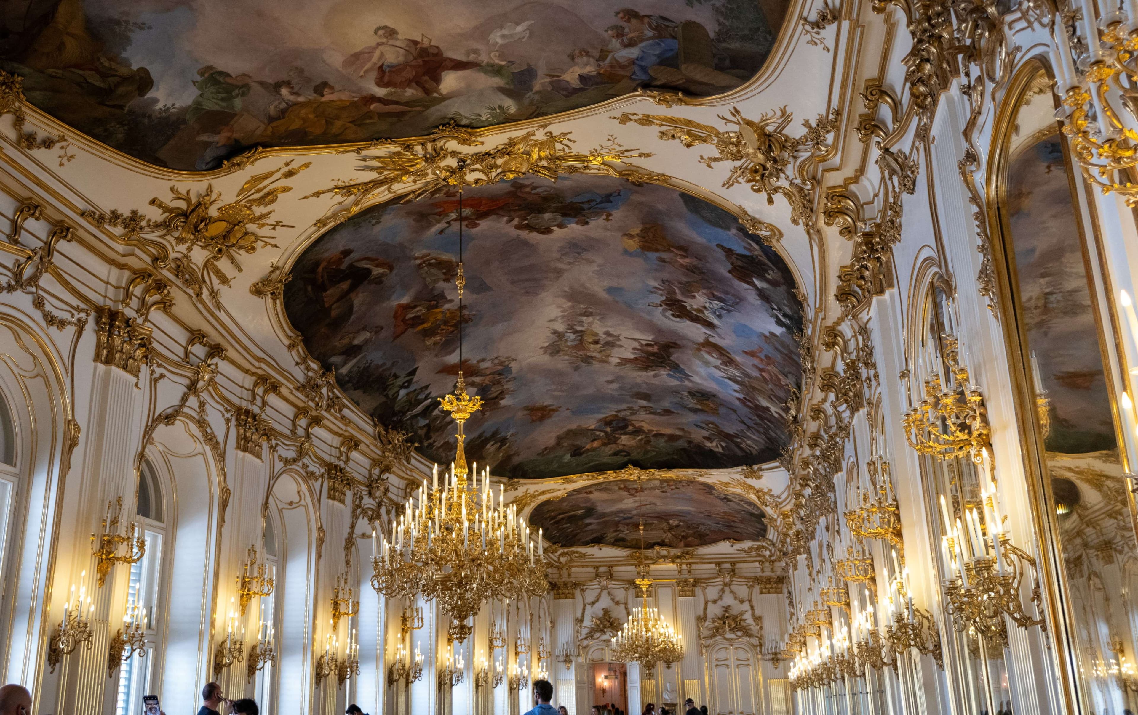 There are many people walking in a large room with a ceiling painted with paintings.