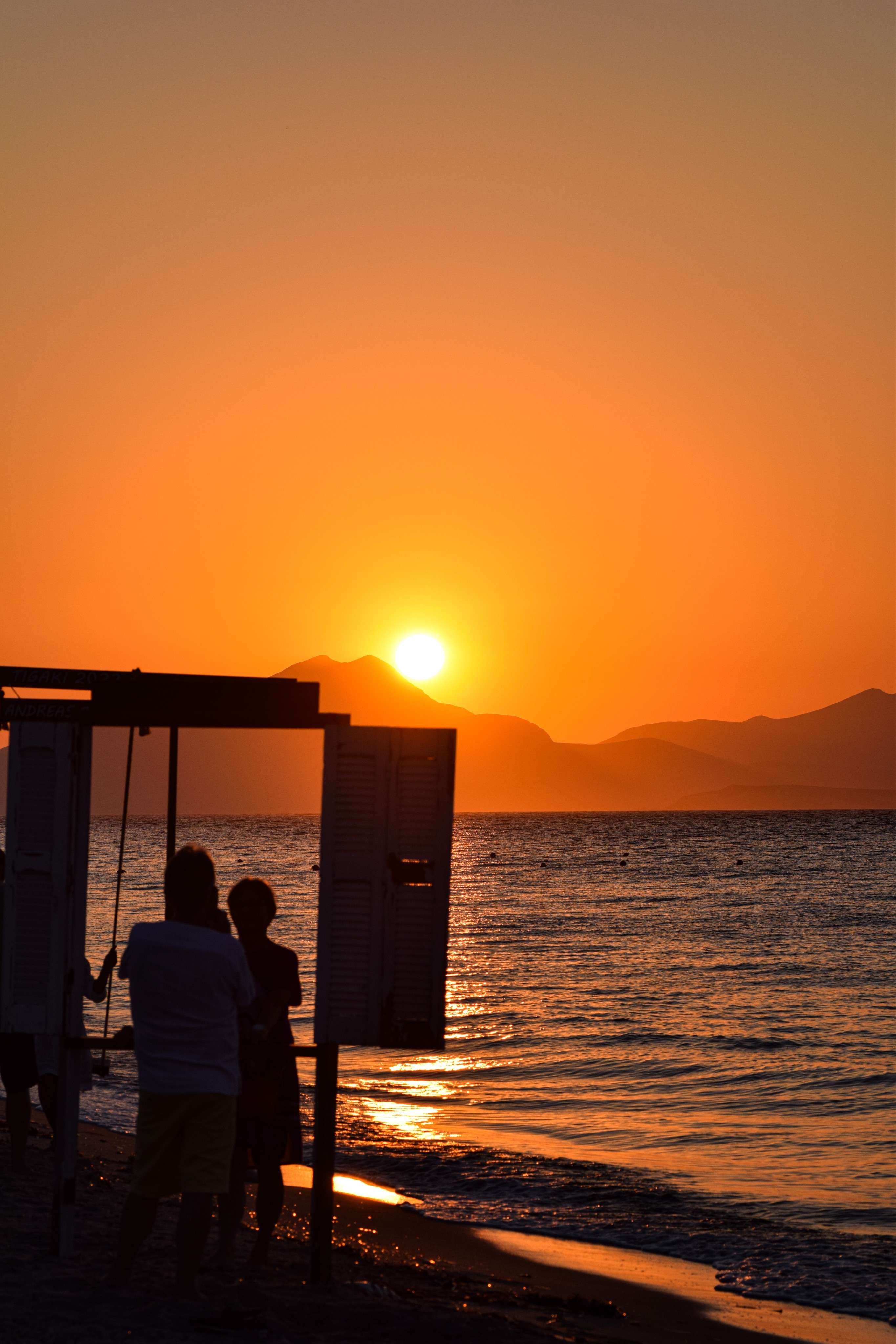 Sunset on the beach with people standing on the sand and a boat in the water.